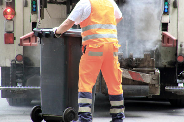 Auf dem Bild schiebt ein Mitarbeiter der Abfallentsorgung in orange-farbener Arbeitsbekleidung eine schwarze Mülltonne Richtung Entsorgungsfahrzeug.