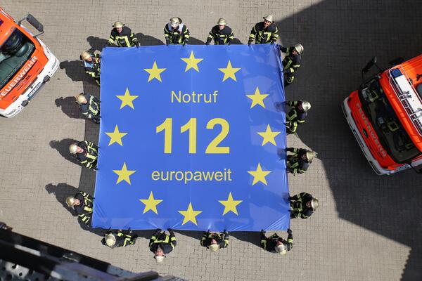 Bild vergrößern: Auf dem Bild ist das Logo des Europäischen Tages des Notrufs am 11.2.: zu sehen. Es halten mehrere Feuerwehrmänner ein großes blaues Laken, auf dem steht: "Notruf 112 europaweit". Dies macht auf die europaweit gültige Notrufnummer 112 aufmerksam.