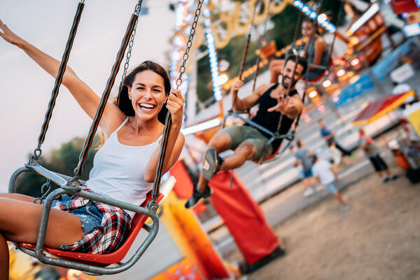 Bild vergrößern: Dargestellt sind lachende junge Leute an einem warmen Tag in einem Kettenkarussell. Im Hintergrund ist ein Ausschnitt eines Riesenrads zu sehen.