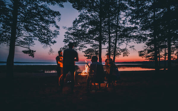 Bild vergrößern: Das Bild zeigt eine kleine Gruppe von Personen an einem See am Abend. In der Mitte der Gruppe befindet sich ein kleines Lagerfeuer