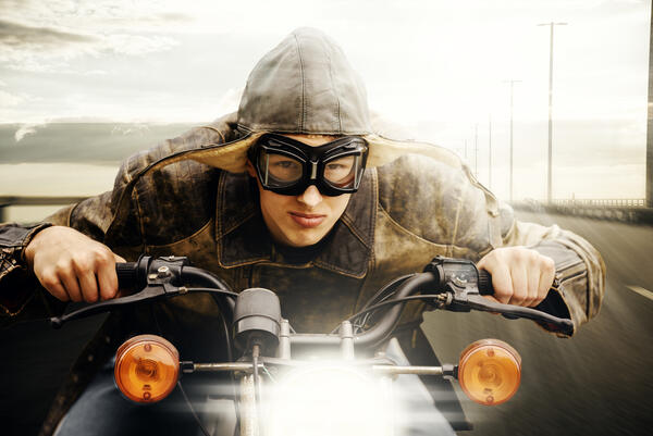 Bild vergrößern: Das Bild zeigt einen jungen Mann mit einer Fliegerbrille und einer Pilotenfliegermütze in Leder der sich weit auf den Lenker seines fahrenden Motorrads lehnt.
