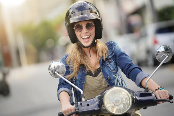 Bild vergrößern: Das Bild zeigt eine lachende junge Frau auf einem Roller. Sie trägt einen schwarzen Helm und eine runde Sonnenbrille mit dunkel getönten Scheiben.