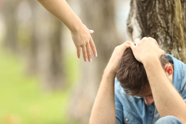 Bild vergrößern: Das Bild zeigt einen jungen Mann der hockend an einen Baum gelehnt ist und die Hände über den gesenkten Kopf hat. Daneben sieht man eine Hand die ihm entgegen gestreckt wird.