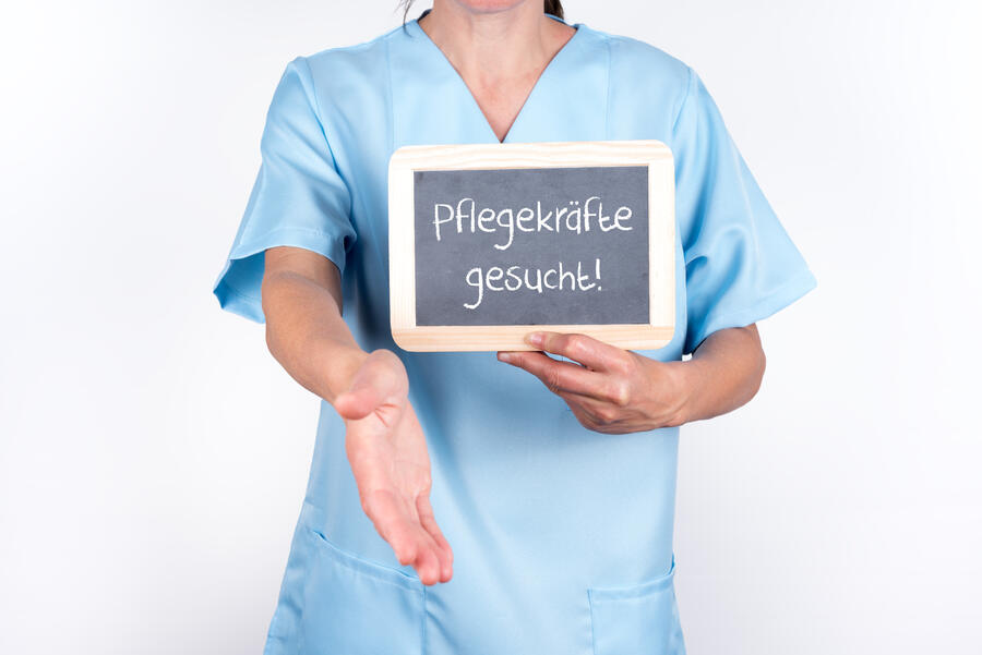 Bild vergrößern: Eine Mitarbeiterin des Pflegepersonals reicht die rechte Hand nach vorne und hält in der linken Hand ein Schild mit der Aufschrift "Pflegekräfte gesucht!"