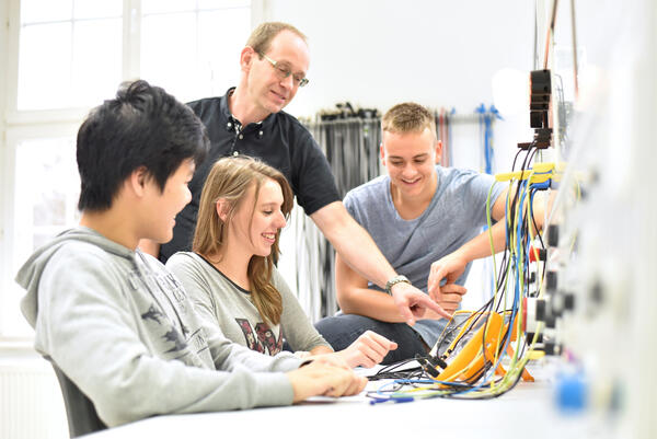 Auf dem Bild sieht man einen Lehrer, der drei Schülern Elektrotechnik erklärt.