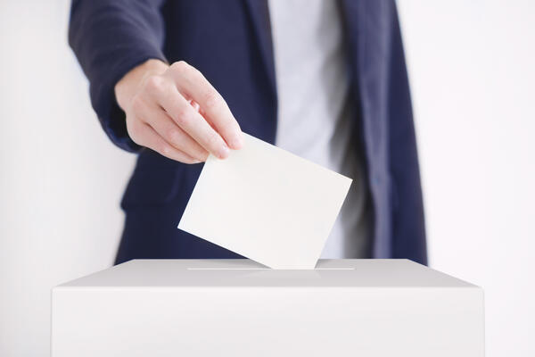 Das Bild zeigt, wie eine männliche Person mit blauem Jackett einen zusammengefalteten Wahlscheins in die Wahlurne steckt. 