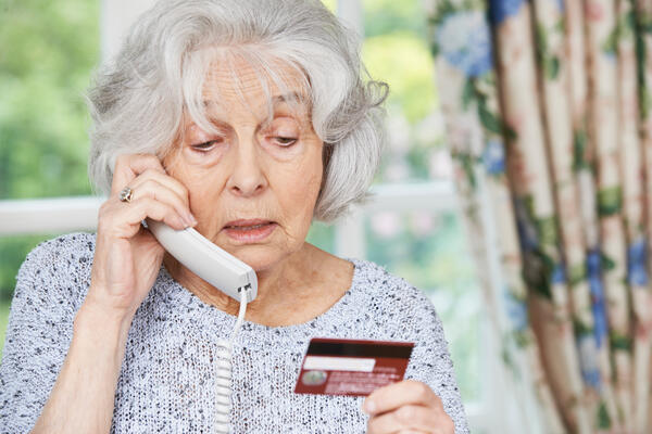 Das Bild zeigt eine ältere Frau, die gerade telefoniert und dabei ihre Bankkarte in der Hand hält. Offenbar sagt sie dem Gegenüber ihre Kontonummer an. Dies sollte generell nie geschehen. 