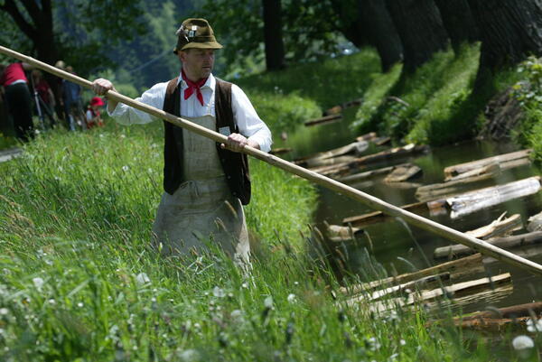 Das Bild zeigt einen Flößer mit seiner Flößstange, wie er an einem Bach Holzscheite entlang schiebt. 