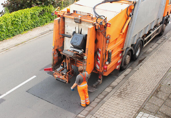 Bild vergrößern: Abfallbehälter wird an einem orangefarbenen Müllfahrzeug gekippt.