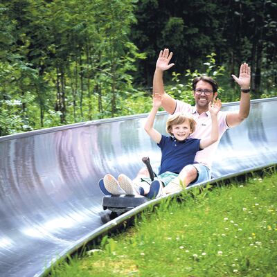 Bild vergrößern: Das Bild zeigt Vater und Sohn, wie sie auf der Sommerrodelbahn mit gehobenen Händen nach unten fahren.