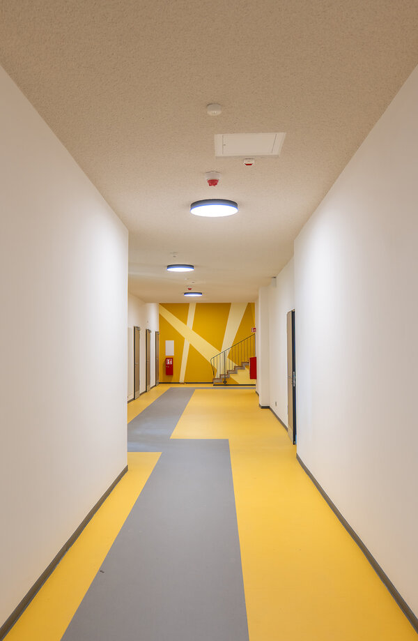 Das Bild zeigt ein neu saniertes Erdgeschoss einer Schule. Es hat einen gelben Flur mir einem grauen Streifen und ist sehr modern gehalten.
