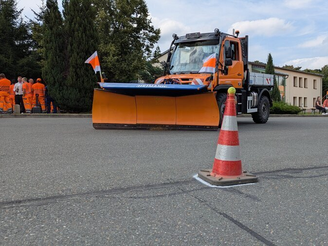 Ein orangener Unimog mit Schaufel steht hinter einem Kegel.