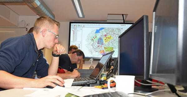 Bild vergrößern: Das Bild zeigt Teilnehmer der Übung des Brand- und Katastrophenschutzes bei konzentriertem Arbeiten an ihren Computern. Im Hintergrund befindet sich eine Karte des Vogtlandkreises.