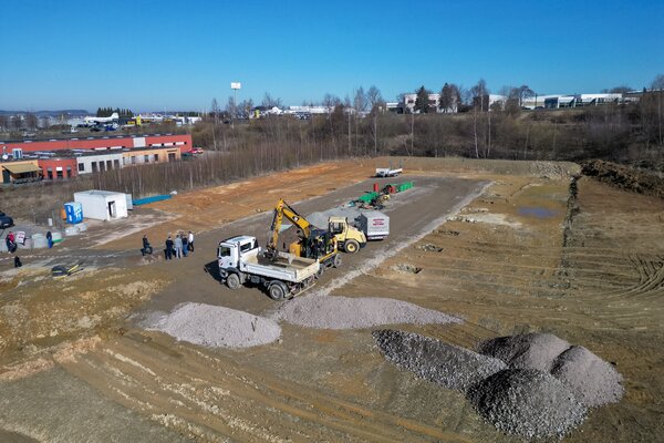 Bild vergr&ouml;&szlig;ern: Luftbild der Baustelle in Reichenbach: Auf dem Gel&auml;nde stehen Bagger, Lkw und Baumaschinen, daneben sind mehrere Menschen beim Spatenstich zu sehen. Im Hintergrund liegen Gewerbebauten und eine leicht h&uuml;gelige Landschaft unter blauem Himmel.