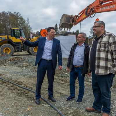 Bild vergr&ouml;&szlig;ern: Landrat Thomas Hennig, Adorfs B&uuml;rgermeister Rico Schmidt und Edeka-Kaufmann Marcel Schuldes stehen auf dem unbefestigten Baustellengel&auml;nde. Landrat zeigt mit dem Arm &uuml;ber das Areal, w&auml;hrend die beiden anderen zuh&ouml;ren. Hinter ihnen stehen ein gro&szlig;er Bagger, ein Radlader und weitere Baumaschinen.
