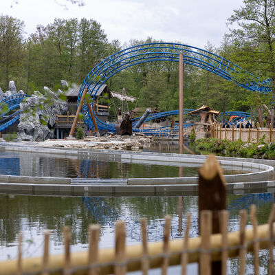 Bild vergr&ouml;&szlig;ern: &Uuml;bersicht der neuen Wasser-Achterbahn &raquo;D&oslash;nnervind": Blaue Schienen f&uuml;hren in hohem Bogen &uuml;ber das Wasserbecken, dahinter Felsformationen und Holzbauten.
