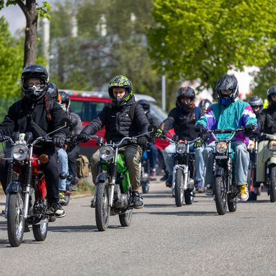 Bild vergr&ouml;&szlig;ern: Eine Gruppe Mopedfahrer mit Helmen sitzt auf historischen Simson-Mopeds und f&auml;hrt nebeneinander auf einer Stra&szlig;e. Am Stra&szlig;enrand stehen weitere Fahrzeuge.