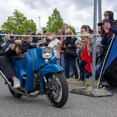 Bild vergr&ouml;&szlig;ern: Ein Fahrer liegt flach auf der Sitzbank einer blauen Simson und unterquert eine Limbo-Stange. Zuschauer verfolgen den Wettbewerb, einige filmen mit ihren Handys.