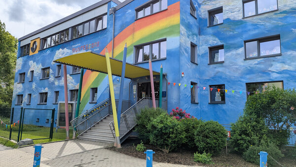 Bild vergr&ouml;&szlig;ern: Ein blaues Haus, auf dem ein Regenbogen zu sehen ist. Der Eingangsbereich ist mit gro�en, bunten St�tzpfeilern in Stiftform umrahmt.