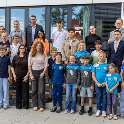 Bild vergr&ouml;&szlig;ern: Eine gr&ouml;&szlig;ere Gruppe aus Kindern, Jugendlichen und Erwachsenen steht vor einer Glasfassade. Mehrere Kinder tragen blaue Sportshirts, die Erwachsenen stehen dahinter und daneben.