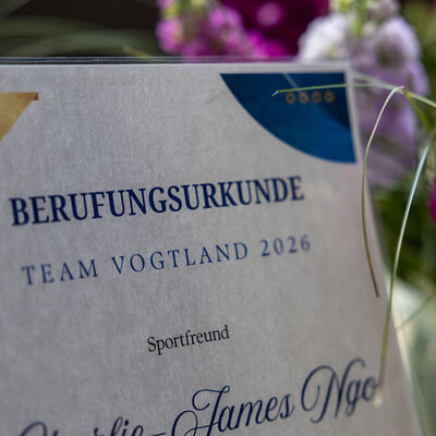 Bild vergr&ouml;&szlig;ern: Nahaufnahme einer Urkunde mit der Aufschrift &acirc;��Berufungsurkunde Team Vogtland 2026&acirc;��. Im Hintergrund sind unscharf Blumen und der Veranstaltungsraum zu erkennen.