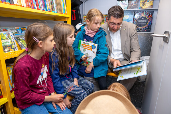 Bild vergr&ouml;&szlig;ern: Landrat Thomas Hennig sitzt mit mehreren Grundschulkindern im Kreisb&uuml;cherbus und schaut gemeinsam mit ihnen in ein aufgeschlagenes Kinderbuch. Links und im Hintergrund sind B&uuml;cherregale zu sehen.