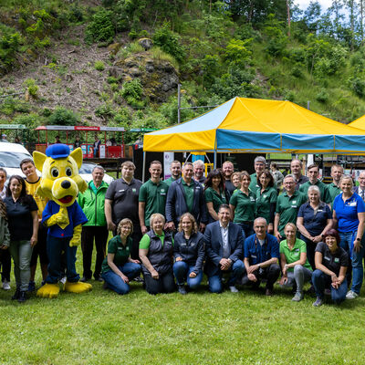 Bild vergr&ouml;&szlig;ern: Eine gro&szlig;e Gruppe von Erwachsenen steht und kniet f&uuml;r ein Gruppenfoto auf einer Wiese. Neben ihnen steht eine gelbe Maskottchenfigur. Im Hintergrund sind Zelte, ein kleiner Zug der Parkeisenbahn und St&auml;nde des Verkehrstages zu sehen.