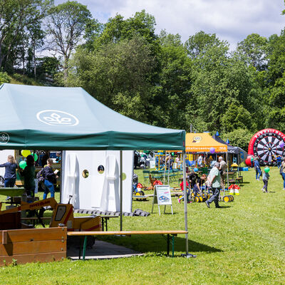 Bild vergr&ouml;&szlig;ern: Blick &uuml;ber eine Wiese mit mehreren Zelten, St&auml;nden und Spielangeboten. Familien laufen zwischen den Stationen umher. Im Hintergrund sind B&auml;ume, ein gro&szlig;es Fu&szlig;ball-Dart-Spiel und weitere Aktionsfl&auml;chen zu sehen.