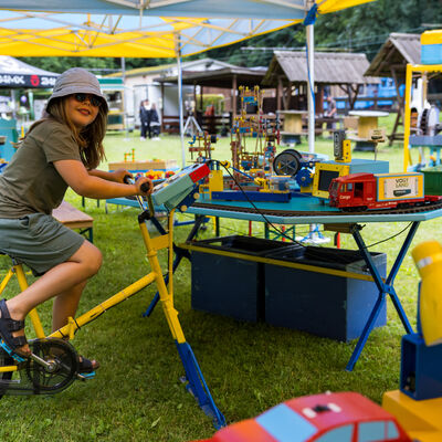 Bild vergr&ouml;&szlig;ern: Ein Kind mit Sonnenbrille sitzt auf einem feststehenden gelben Fahrrad und tritt in die Pedale. Vor ihm steht ein buntes Modell mit kleinen Fahrzeugen und technischen Elementen. Die Szene spielt unter einem Zelt auf einer Wiese.
