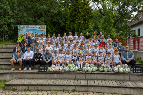 Bild vergr&ouml;&szlig;ern: Gruppenfoto der ersten Trainingswoche: Kinder und Jugendliche in wei&szlig;en Fu&szlig;balltrikots sitzen und stehen mit Trainern und G&auml;sten auf einer Treppe im Waldpark Gr&uuml;nheide. Vor ihnen liegen mehrere Fu&szlig;b&auml;lle.