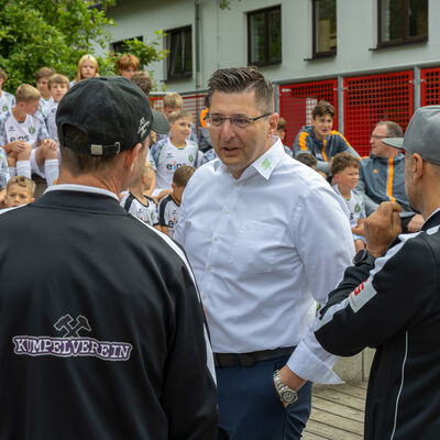 Bild vergr&ouml;&szlig;ern: Der Landrat spricht mit zwei Trainern. Dahinter sitzen viele Kinder in Fu&szlig;balltrikots auf einer Trib&uuml;ne und warten auf den Beginn des Trainings.