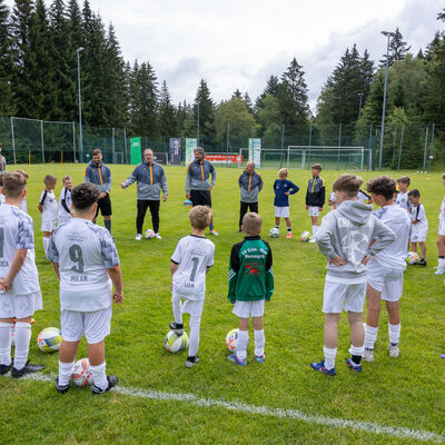 Bild vergr&ouml;&szlig;ern: Kinder und Jugendliche in wei&szlig;en Fu&szlig;balltrikots stehen im Kreis auf einem Rasenplatz. Vor ihnen liegen Fu&szlig;b&auml;lle. Mehrere Trainer sprechen zur Gruppe. Rund um den Platz stehen Tore, Trainingsstangen und Wald.