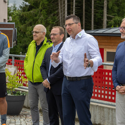 Bild vergr&ouml;&szlig;ern: Der Landrat spricht bei der Er&ouml;ffnung der Fu&szlig;ballschule im Waldpark Gr&uuml;nheide. Neben ihm stehen mehrere Vertreter und Trainer. Im Hintergrund ist Wald zu sehen.
