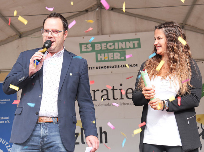 Michael Groh steht auf einer B&uuml;hne und verk&uuml;ndet den Namen der Kinderstadt. Es fliegt Konfetti.