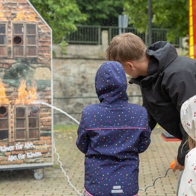 Bild vergr&ouml;&szlig;ern: Zwei Kinder richten gemeinsam mit einem Betreuer einen Wasserstrahl auf die Darstellung eines brennenden Hauses. Im Hintergrund steht ein rotes Feuerwehrfahrzeug.