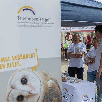 Bild vergr&ouml;&szlig;ern: Landrat Thomas Hennig spricht mit zwei Ehrenamtlichen am Stand der Telefonseelsorge. Im Vordergrund steht ein Banner mit einer Eule.