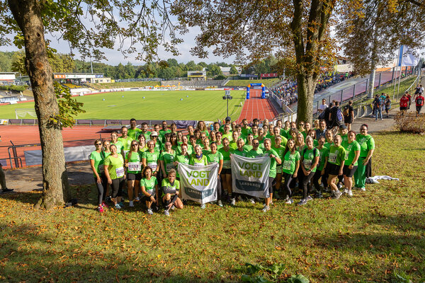 Eine gro&szlig;e Gruppe von Mitarbeiterinnen und Mitarbeitern des Vogtlandkreises steht in gr&uuml;nen Laufshirts vor dem Vogtlandstadion. In der ersten Reihe werden zwei Fahnen mit der Aufschrift &raquo;Vogtland Landkreis&laquo; gehalten.
