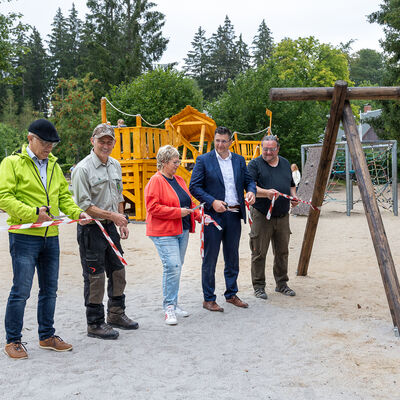 Bild vergr&ouml;&szlig;ern: F&uuml;nf Personen durchschneiden gemeinsam ein rot-wei&szlig;es Band auf einem Spielplatz. Im Hintergrund befinden sich h&ouml;lzerne Kletter- und Spielelemente.