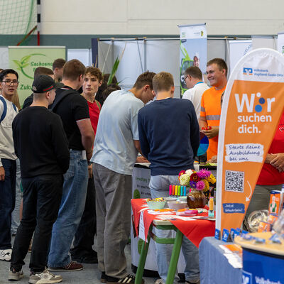 Bild vergr&ouml;&szlig;ern: Mehrere Jugendliche stehen vor verschiedenen Messest&auml;nden in der Sporthalle und sprechen mit Ausstellern. Roll-ups und Informationsmaterialien sind rund um die St&auml;nde aufgebaut.