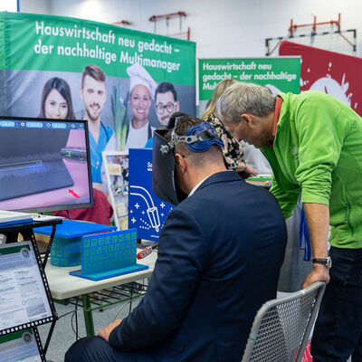 Bild vergr&ouml;&szlig;ern: Landrat Thomas Hennig sitzt an einem Messestand und tr&auml;gt eine VR-Brille. Ein Aussteller steht neben ihm und begleitet die virtuelle Schwei&szlig;&uuml;bung.