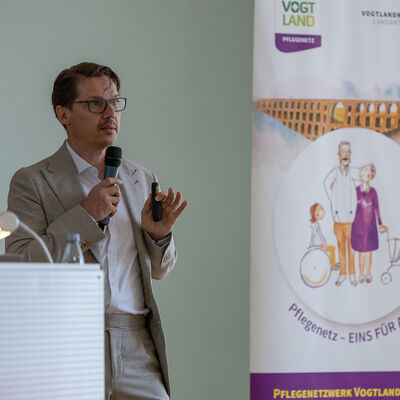 Bild vergr&ouml;&szlig;ern: Markus Seibt vom Landesverband f&uuml;r Hospizarbeit und Palliativmedizin Sachsen spricht mit einem Mikrofon vor dem Publikum und h&auml;lt eine Pr&auml;sentationsfernbedienung in der Hand. Neben ihm steht ein Banner des Pflegenetzwerkes Vogtlandkreis.