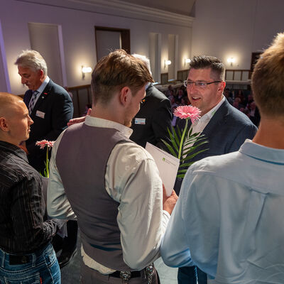 Bild vergr&ouml;&szlig;ern: Landrat Thomas Hennig steht auf der B&uuml;hne zwischen mehreren jungen Handwerkern und gratuliert einem Absolventen. Einige halten Blumen und Urkunden in den H&auml;nden, im Hintergrund sitzt das Publikum.