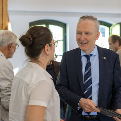 Bild vergr&ouml;&szlig;ern: Dr. Axel Steinbach im dunklen Anzug spricht in einem hellen Veranstaltungsraum mit Dr. Christine Heuck. Im Hintergrund stehen weitere Teilnehmerinnen und Teilnehmer des Jahrestreffens.