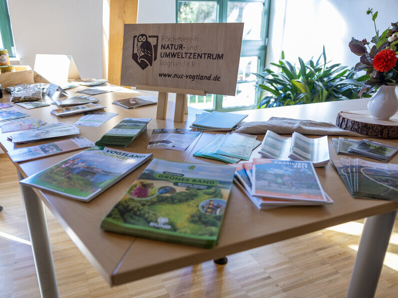 Bild vergr&ouml;&szlig;ern: Auf einem Tisch liegen Brosch&uuml;ren und Informationsbl&auml;tter zu Natur- und Umweltthemen. In der Mitte steht ein Holzschild des Natur- und Umweltzentrums Vogtland.