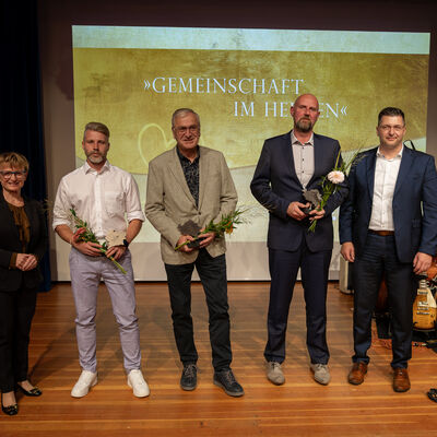 Bild vergr&ouml;&szlig;ern: Gruppenfoto der Kategorie &raquo;Sport&laquo; mit drei ausgezeichneten M&auml;nnern, Blumen und Ehrenamtspokalen auf der B&uuml;hne. Im Hintergrund ist das Motto &raquo;Gemeinschaft im Herzen&laquo; zu sehen.