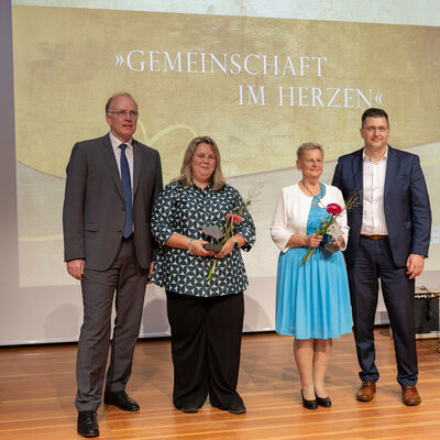 Bild vergr&ouml;&szlig;ern: Vier Personen stehen vor der goldfarbenen B&uuml;hnenwand mit dem Schriftzug &raquo;Gemeinschaft im Herzen&laquo;. In der Mitte halten zwei ausgezeichnete Frauen Pokale und Blumen, links steht Marco Fuchs, rechts Landrat Thomas Hennig.