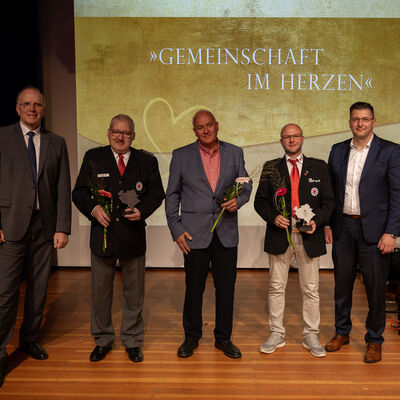 Bild vergr&ouml;&szlig;ern: F&uuml;nf M&auml;nner stehen f&uuml;r ein Gruppenfoto auf der B&uuml;hne. Drei Geehrte in der Mitte halten Blumen und Ehrenamtspokale, zwei von ihnen tragen DRK-Uniform. Links steht Marco Fuchs, rechts Landrat Thomas Hennig.