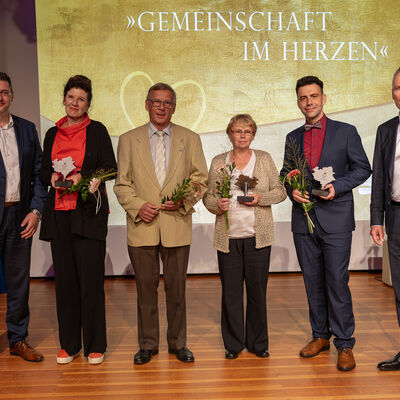 Bild vergr&ouml;&szlig;ern: Sechs Personen stehen nebeneinander auf der B&uuml;hne. Vier Geehrte in der Mitte halten Blumen und Ehrenamtspokale, zwei weitere Personen flankieren die Gruppe. Hinter ihnen ist das Motto &raquo;Gemeinschaft im Herzen&laquo; zu sehen.
