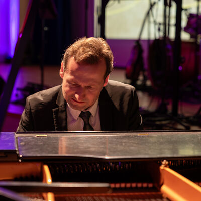 Bild vergr&ouml;&szlig;ern: Ein Pianist im dunklen Anzug sitzt konzentriert am ge&ouml;ffneten Fl&uuml;gel. Im Hintergrund ist die farbig beleuchtete B&uuml;hne des K&ouml;niglichen Kurhauses zu sehen.