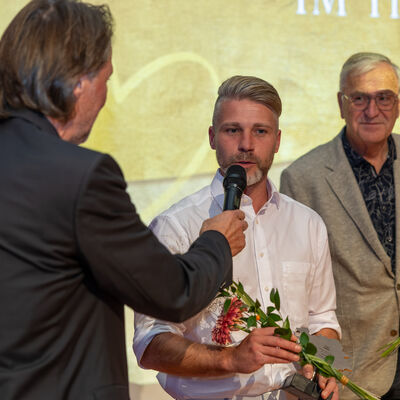 Bild vergr&ouml;&szlig;ern: Philipp Funke steht mit Blumen und Ehrenamtspokal auf der B&uuml;hne und beantwortet im Interview Fragen. Neben ihm ist ein weiterer ausgezeichneter Mann zu sehen.